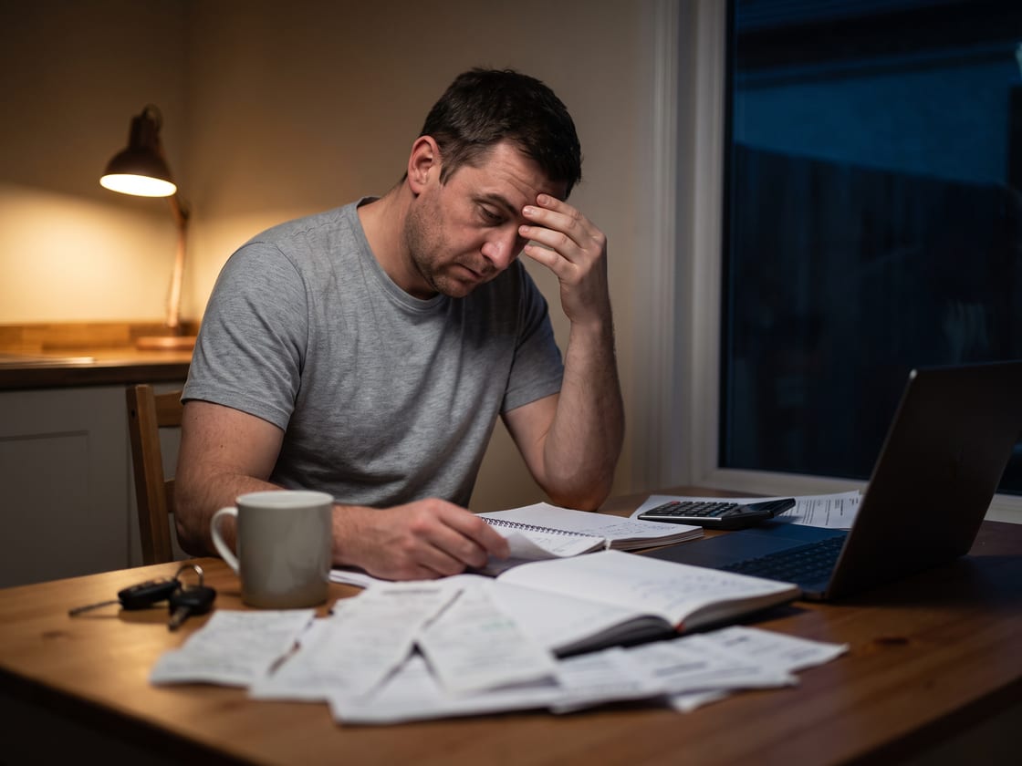 A business owner at a kitchen table late at night, surrounded by paper invoices, a notebook and a calculator