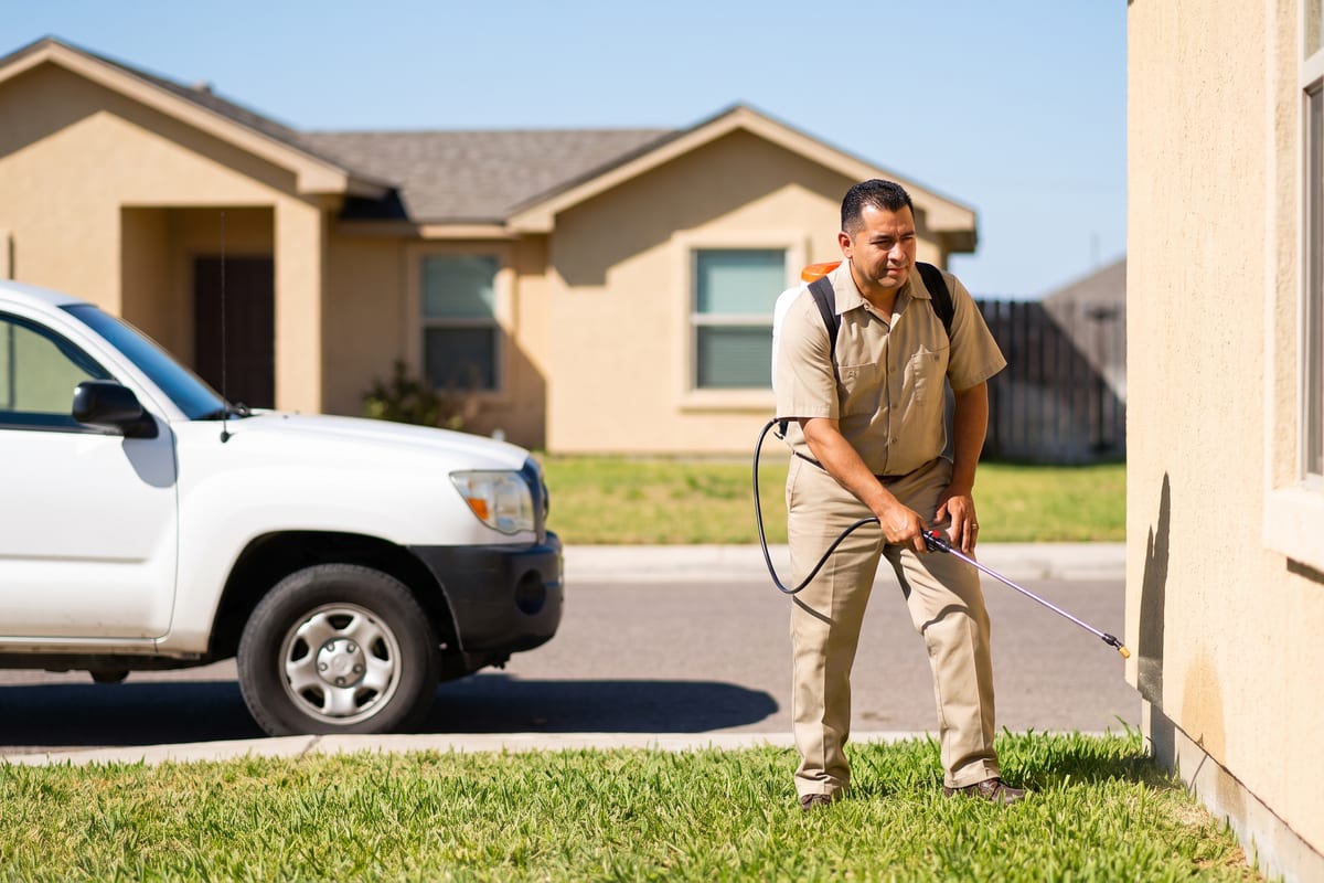 Pest control technician in a khaki uniform treating along the exterior foundation of a home with a backpack sprayer