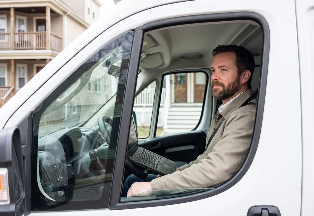 Business owner in the cab of a van with a laptop on the steering wheel outside a small apartment building